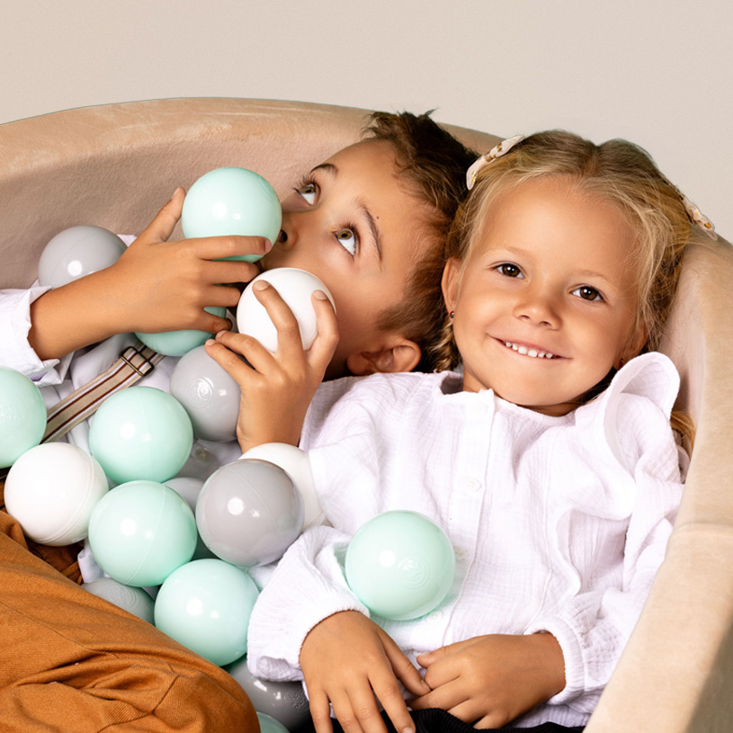 To børn leger i kuglebad med hvide og mintgrønne bolde, en af dem smiler bredt.